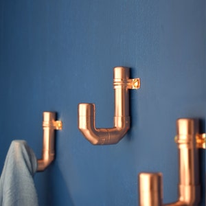 May include: Three copper pipe coat hooks mounted on a blue wall. The hooks are in the shape of a 'J' and are made of copper pipe.