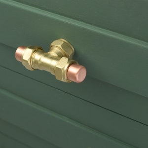 May include: A brass and copper T-shaped pipe fitting, mounted on a green surface. The fitting has a polished finish, with hexagonal nuts and copper end caps. The background is a dark green, with visible wood grain. The fitting is a plumbing component.