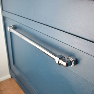 May include: A chrome-plated metal drawer pull with a pipe-style design. The pull is mounted on a blue painted cabinet door.