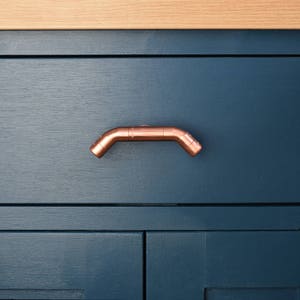 May include: A copper drawer pull in the shape of a curved pipe. The pull is mounted on a dark blue cabinet door.