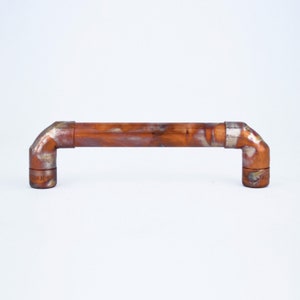 Peut inclure: Une poignée de porte en cuivre avec une finition rustique. La poignée est en forme d'angle de 90 degrés et a une bande argentée autour du centre.