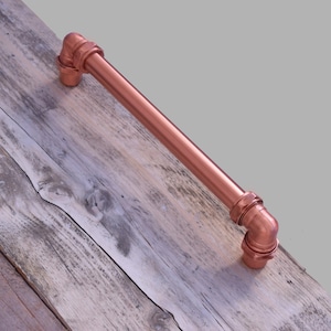 May include: A copper pipe drawer pull with a 90-degree bend. The pull is on a weathered wooden surface.