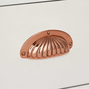 May include: A copper shell-shaped drawer pull with a smooth, shiny finish. The pull has a slightly curved shape and is designed to be used on cabinets or drawers.