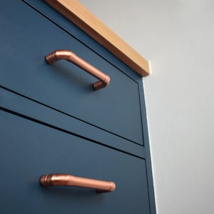 May include: Two drawers with copper handles on a dark blue cabinet. The cabinet is made of wood and has a light wood top.