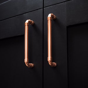 May include: Two copper cabinet door pulls with a pipe design. The pulls are mounted on a black cabinet door.