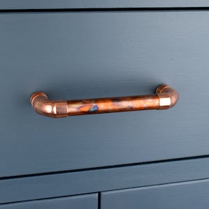 Könnte beinhalten: Ein Schubladenzug aus Kupferrohr mit leicht oxidierter Oberfläche. Der Zug ist an einer dunkelblauen Schranklade montiert.