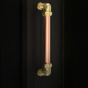 May include: A copper pipe door handle with brass fittings. The handle is mounted on a black wall.