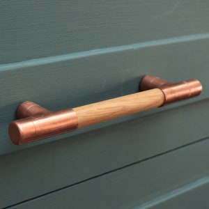 May include: A copper and wood cabinet pull with a rectangular shape. The pull has a wooden centre and copper ends.