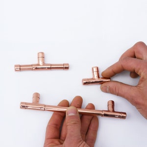 May include: Three copper pipe drawer pulls. The pulls are in the shape of a T with a short horizontal bar. The pulls are shown on a white surface.