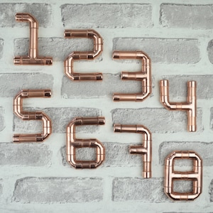 May include: Copper pipe house numbers 1 through 8, mounted on a white brick wall. The numbers are made of copper pipe and are in a simple, modern style.