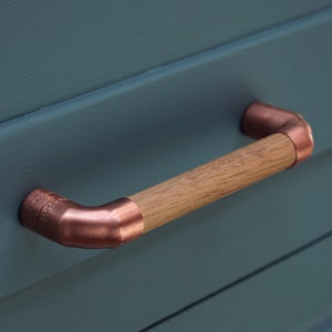 Puede incluir: Un tirador de armario de cobre y madera con forma rectangular redondeada. El tirador está hecho de tubería de cobre con un mango de madera.