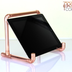 May include: A copper pipe tablet stand with a black tablet. The stand is made of copper pipes and fittings, with the tablet resting at an angle. The stand is designed to hold a tablet securely. The logo "Proper Copper" is in the top right corner.
