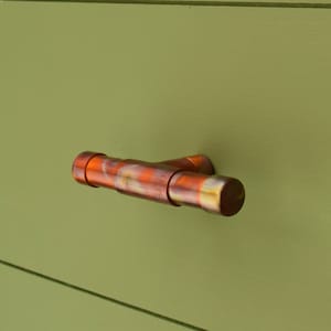 以下が含まれることがあります： 緑色の塗装面に取り付けられた、酸化して斑点のある仕上げの銅製のT字型のキャビネットプルです。