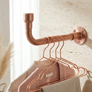 May include: A copper-colored wall-mounted clothing rack with a pipe design. The rack holds several copper-colored wire hangers with clothing. The rack is mounted on a textured, light-colored wall. The design is industrial and modern.