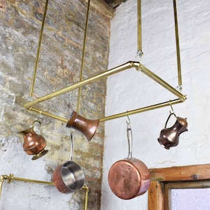 May include: Brass pot rack with copper pots and jugs hanging from it. The rack is mounted to the ceiling and is made of square tubing.