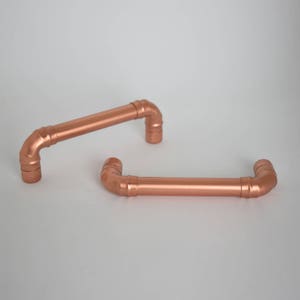 May include: Two copper pipe drawer pulls. The pulls are in the shape of a 90-degree angle with a straight section in the middle.