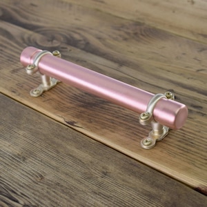 May include: A rose gold drawer pull with a cylindrical shape and silver accents. The pull is mounted on a wooden surface.