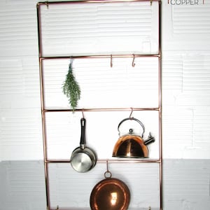 Può includere: Un portaoggetti in tubo di rame con quattro ripiani e ganci per appendere utensili da cucina. Un bollitore in rame, una pentola e una ciotola in rame sono appesi ai ganci. Un mazzo di erbe è appeso al ripiano superiore. Il portaoggetti è montato su una parete di mattoni bianchi. Il testo "proper copper" è stampato nell'angolo in alto a destra dell'immagine.