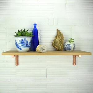 Könnte beinhalten: Ein Holzregal mit Kupferkonsolen, auf dem eine blaue Glasflasche, ein weißer Keramiktopf mit blau-weißem Muster, ein goldfarbener Engelsflügel und ein weißer Keramiktopf mit blau-weißem Muster stehen. Das Regal ist an einer weißen Backsteinwand montiert.