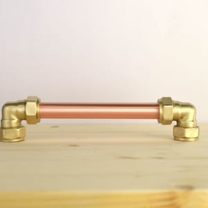 Könnte beinhalten: Ein Schubladenzug aus Kupferrohr mit goldfarbenen Beschlägen. Der Zug ist auf einer hellholzfarbenen Oberfläche montiert.