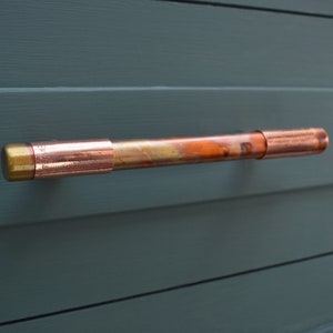 May include: A copper pipe door handle with a gold end cap. The handle is mounted on a dark green wall.