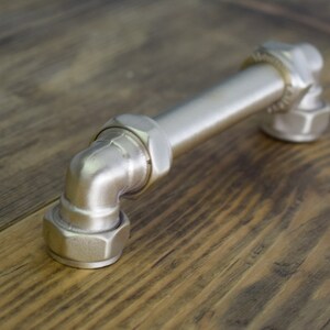 May include: A silver metal pipe fitting door handle with a 90-degree bend. The handle is made of industrial pipe and has a polished finish.