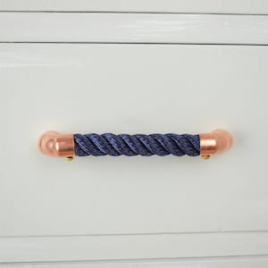 May include: A dark blue rope drawer pull with copper end caps. The pull is mounted on a white cabinet door.