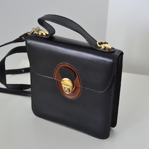 May include: A structured black leather handbag featuring a top handle and a detachable shoulder strap. The front flap is secured with a gold-tone metal push-lock clasp set against a circular brown leather accent patch with black trim.