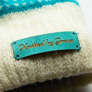 Op de afbeelding: Een witte gebreide muts met een blauw-wit gestreept patroon. Een turquoise lederen label is op de muts genaaid met de tekst "Knitted by Grace" in gouden letters.