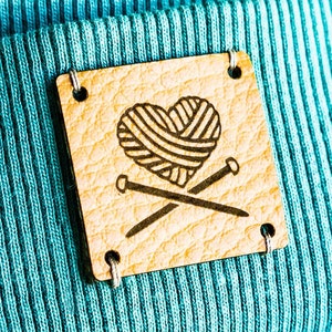 Könnte beinhalten: Eine türkisblaue Strickmütze mit einem braunen Lederflicken, der ein Herz mit einem Wollknäuel und zwei gekreuzten Stricknadeln zeigt.
