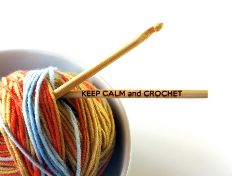 May include: A ball of yarn in orange, blue, and yellow is in a white bowl. A wooden crochet hook with the text "KEEP CALM and CROCHET" is resting on top of the yarn.
