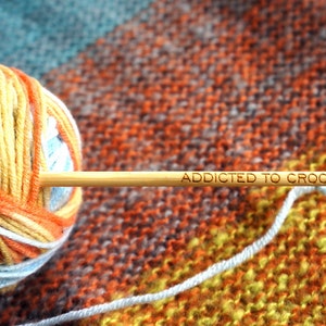 Może przedstawiać: Kulka pomarańczowej, ż&oacute;łtej i białej wł&oacute;czki z drewnianym haczykiem do szydełkowania, na kt&oacute;rym napisano "Addicted to Crochet".
