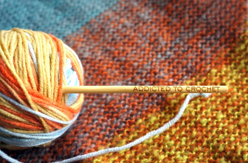 Może przedstawiać: Kulka pomarańczowej, ż&oacute;łtej i białej wł&oacute;czki z drewnianym haczykiem do szydełkowania, na kt&oacute;rym napisano "Addicted to Crochet".