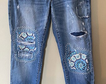 May include: Blue denim jeans with embroidered patches on the knees. The patches are a blue and white paisley design. The jeans have distressed details and are a straight leg style.