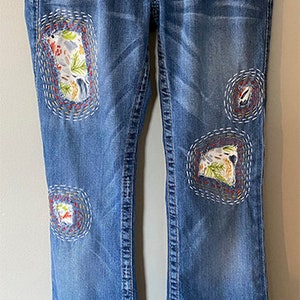 May include: Blue denim jeans with decorative patches. The patches feature embroidered designs with floral and landscape motifs. The jeans have a classic five-pocket design, a button and zipper closure, and a flared leg opening.