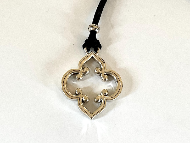 May include: A silver-toned pendant necklace with a four-leaf clover design. The pendant is suspended from a black cord. The necklace is displayed against a white background.