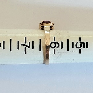 May include: A gold ring with a red gemstone set in a square shape. The ring is on a ruler with markings in inches.