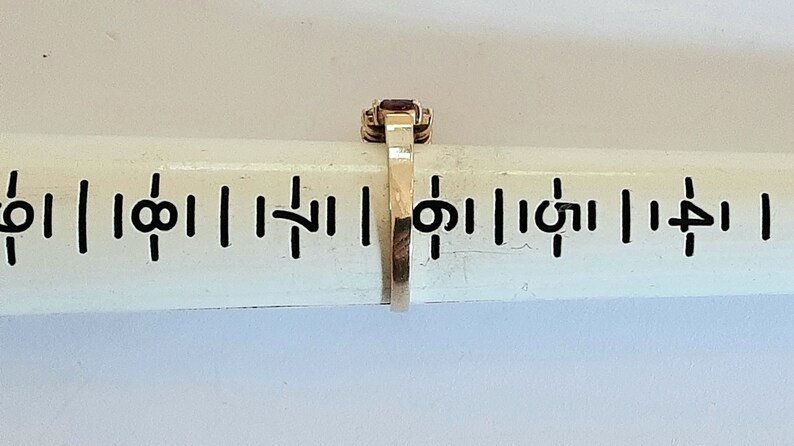 May include: A gold ring with a red gemstone set in a square shape. The ring is on a ruler with markings in inches.