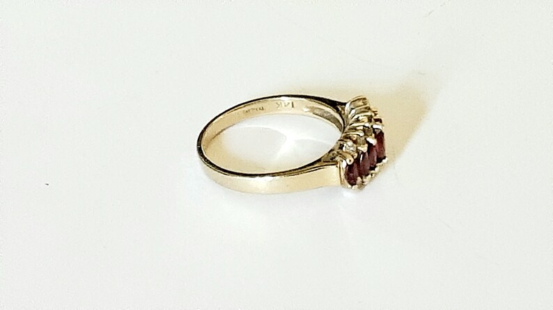 May include: A gold ring with a row of red gemstones and smaller clear gemstones set in the band. The ring is marked "14K" on the inside.