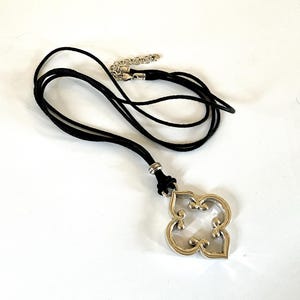 May include: A silver-toned pendant necklace with a black cord. The pendant has a four-leaf clover design with a decorative, openwork pattern. The necklace has an adjustable chain.