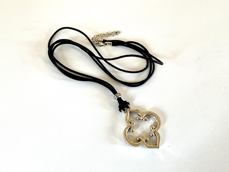 May include: A silver-toned pendant necklace with a black cord. The pendant has a four-leaf clover design with a decorative, openwork pattern. The necklace has an adjustable chain.