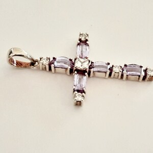 Pu&ograve; includere: Un ciondolo a forma di croce in argento con una pietra centrale trasparente e pietre baguette viola.