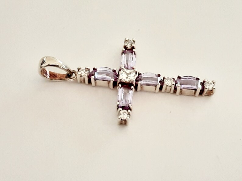 Pu&ograve; includere: Un ciondolo a forma di croce in argento con una pietra centrale trasparente e pietre baguette viola.