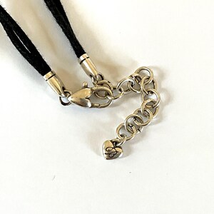 May include: Close-up of a necklace clasp and chain. The necklace features a black cord, silver-toned metal clasps, and a chain with a heart-shaped charm. The chain is made of small, interconnected rings.