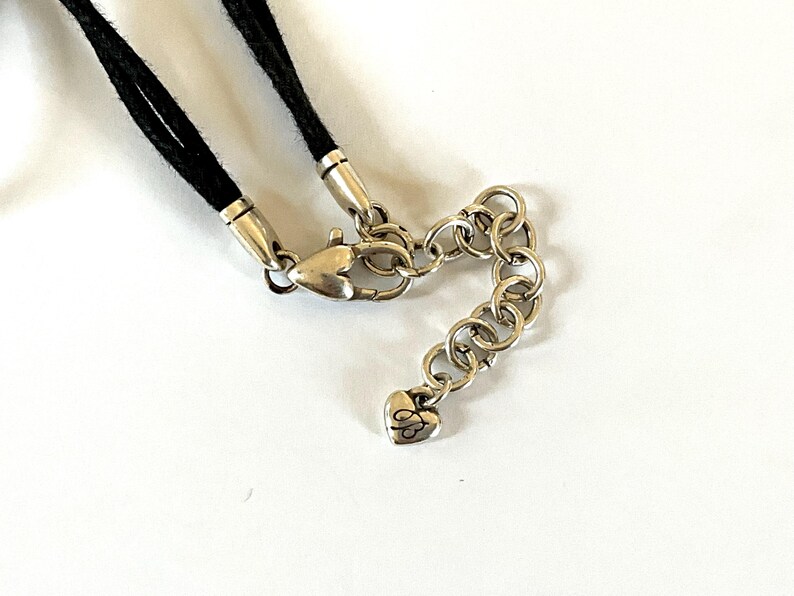 May include: Close-up of a necklace clasp and chain. The necklace features a black cord, silver-toned metal clasps, and a chain with a heart-shaped charm. The chain is made of small, interconnected rings.