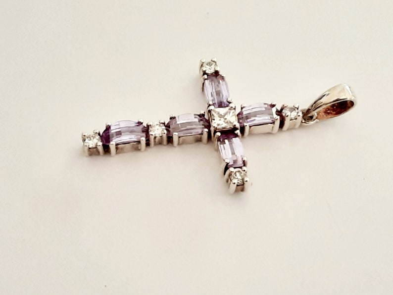 Pu&ograve; includere: Un ciondolo a forma di croce in argento con una grande pietra centrale trasparente e pietre pi&ugrave; piccole di colore viola intorno ai bordi.
