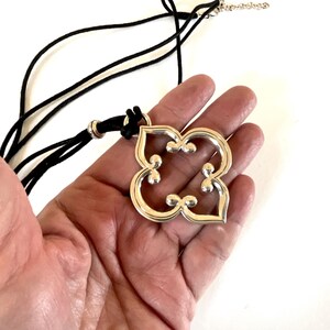 May include: Silver-toned pendant necklace with a black cord. The pendant has a four-petal floral design with small, raised details. The necklace is displayed on a hand against a white background. The pendant's design is symmetrical and modern.