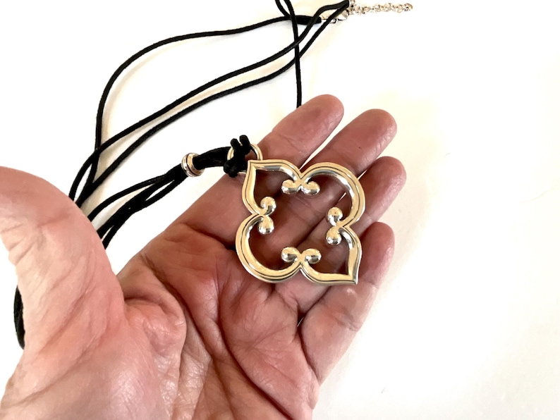 May include: Silver-toned pendant necklace with a black cord. The pendant has a four-petal floral design with small, raised details. The necklace is displayed on a hand against a white background. The pendant's design is symmetrical and modern.