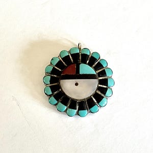 May include: A silver pendant with a circular design. The center features a white and black circle with a small red square. The outer ring has alternating black and turquoise teardrop shapes. A small silver loop is at the top.