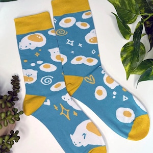 May include: A pair of blue socks with a yellow cuff. The socks feature a white egg design with a yellow yolk and a white cartoon animal.